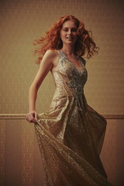 Güzel kızıl saçlı bir kadının portresi lüks altın rengi bir elbiseyle altın duvar kağıdı arka planda poz veriyor. Güzellik, akşam modası.. 