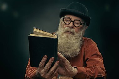 Melon şapkalı ve gözlüklü uzun gri sakallı bilge yaşlı bir adam eski bir kitap okuyor. Siyah arkaplan. 