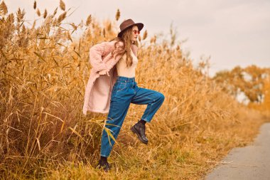 Sonbahar modası. Güz manzarasının arka planında sazlıkların yanında poz veren modaya uygun giyinmiş güzel bir genç kadının tam boy portresi..