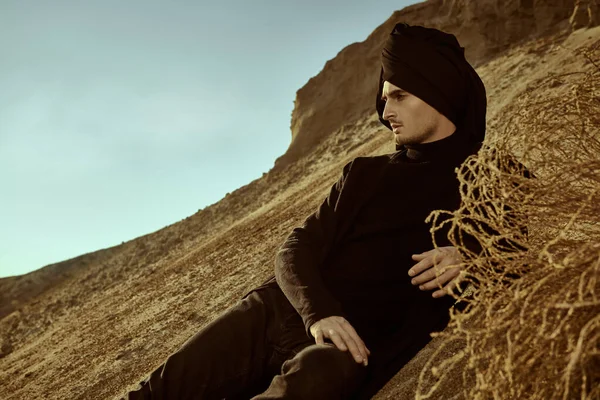 Adventures, the spirit of freedom. Courageous young man in black clothes and black turban on his head sits in a desert and looks pensive forward.