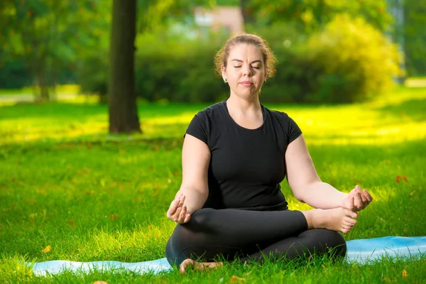 Concentrated woman oversize meditates on the lawn in the park in — Stock Photo, Image