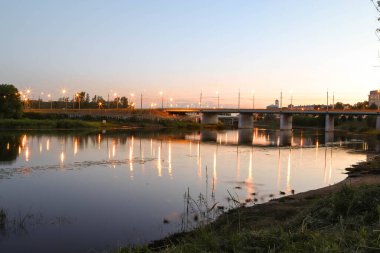 Yaroslavl. Kotorosl nehrinin kıyısında sıcak bir akşam. Kotorosl nehri üzerindeki köprüler gün batımı ve akşam ışıklarının arka planında. Suyu sakinleştirmek için yansıtma. Doğanın yansıması.