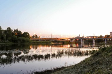 Yaroslavl. Kotorosl nehrinin kıyısında sıcak bir akşam. Kotorosl nehri üzerindeki köprüler gün batımı ve akşam ışıklarının arka planında. Suyu sakinleştirmek için yansıtma. Doğanın yansıması.