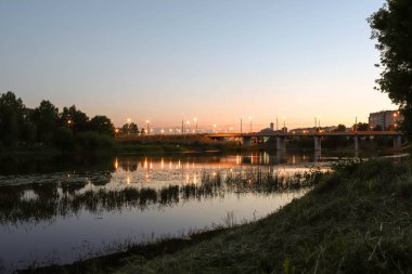 Yaroslavl. Kotorosl nehrinin kıyısında sıcak bir akşam. Kotorosl nehri üzerindeki köprüler gün batımı ve akşam ışıklarının arka planında. Suyu sakinleştirmek için yansıtma. Doğanın yansıması.