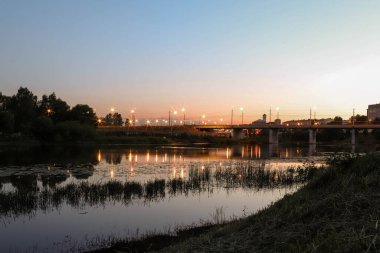 Yaroslavl. Kotorosl nehrinin kıyısında sıcak bir akşam. Kotorosl nehri üzerindeki köprüler gün batımı ve akşam ışıklarının arka planında. Suyu sakinleştirmek için yansıtma. Doğanın yansıması.