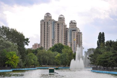 Şehir parkında çeşme. Su sıçraması fıskiyeleri yüksek yukarı yendi. Arka planda, modern tarzda mimarisi ile modern evler. Bir Granit anıt görünümlü-sonunda altın bir yıldız ile bir Stella. 