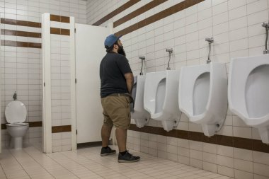 Man with beard urinating in urinal in public toilet clipart