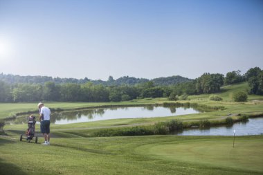 Olgun adam golf oyuncu bir golf sahası ile göl güneşli bir günde onun golf arabasında yakınındaki şapkalı