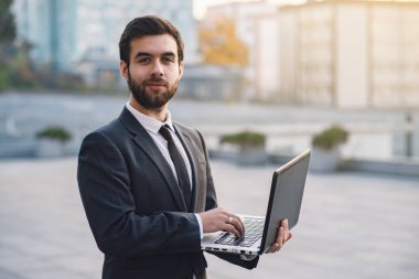Açık havada düz görünümlü bir dizüstü bilgisayar tutan cityscapes bir arka plan üzerinde iş adamı. Kentsel güneşli