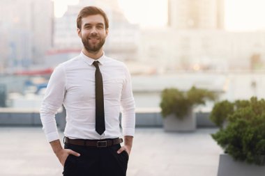 Bir beyaz gömlek ve kravat elleri ceplerinde tutan güneşli bir mutlulukla gülümser şehrin arka planı dostum. Açık havada, arka plan bulanık.