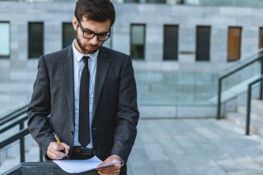 Genç işadamı duran bir takım elbise ve gözlük bir kalem ve boş beyaz kağıt iş merkezi arka plan üzerinde tutar. Açık