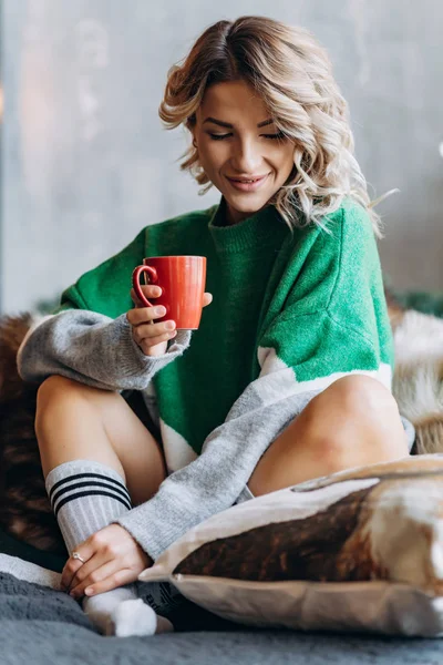 Onun elinde kırmızı bir fincan ile yeşil bir süveterin içinde oturan sarışın gülümseyen bir kadın portresi