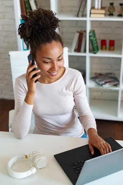 Uśmiechnięta kobieta mulat pracuje na laptopie w biurze i rozmawia na smartfonie. -Obraz — Zdjęcie stockowe