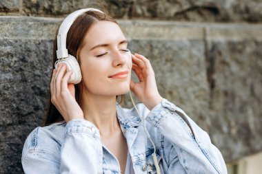 Genç bir öğrenci sokakta, duvara yaslanmış kulaklıkla müzik dinliyor.