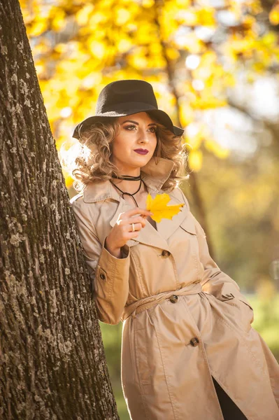 Beautiful blonde woman with cream coat, long legs and black hat in a autumn scene.  Portrait of a very beautiful, elegant and sensual woman with curly hair and sexy legs  posing in autumn park.