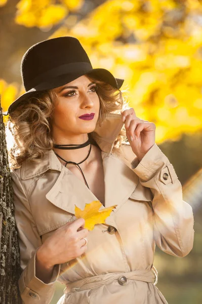 美丽的金发女子与奶油大衣 长腿和黑色的帽子在秋季的场景 一个非常美丽 优雅和感性的女人的肖像 卷曲的头发和性感的腿摆在秋季公园 — 图库照片