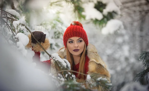 森の冬景色を楽しんでいる茶色の毛皮のケープ付き赤で美しい女性 雪に覆われた木の枝の下でポーズ ブロンドの女の子 明るい寒い日 化粧で周りの雪片と若い女性 — ストック写真