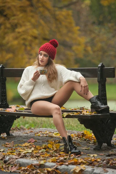 赤い帽子 白いプルオーバーと秋の公園の黒いブーツ 赤い緑の黄色のスリーを持つ美しい若い白人官能的な女性の肖像画 長い足とベンチに座ってコーヒーのカップを持つ女の子 — ストック写真