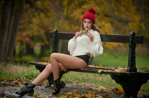 一个美丽的年轻白种人性感女人的肖像与红色帽子 白色拉扯和黑色靴子在秋天公园 红绿黄色三 长腿的女孩和一杯咖啡坐在长凳上 — 图库照片