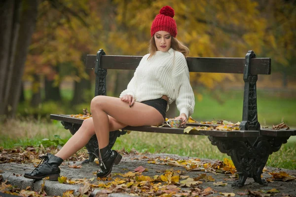 赤い帽子 白いプルオーバーと秋の公園の黒いブーツ 赤い緑の黄色のスリーを持つ美しい若い白人官能的な女性の肖像画 長い足とベンチに座ってコーヒーのカップを持つ女の子 — ストック写真