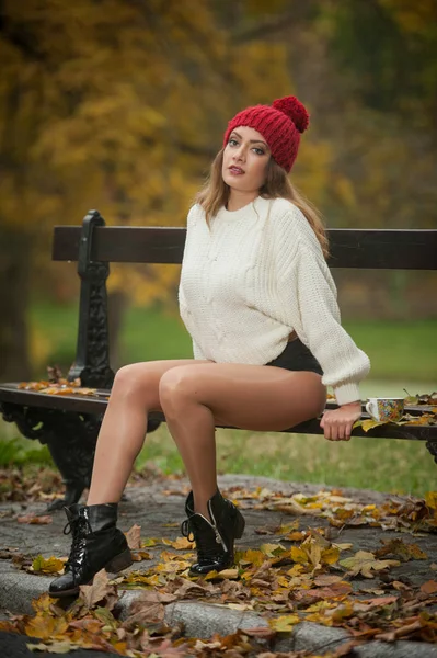 一个美丽的年轻白种人性感女人的肖像与红色帽子 白色拉扯和黑色靴子在秋天公园 红绿黄色三 长腿的女孩和一杯咖啡坐在长凳上 — 图库照片