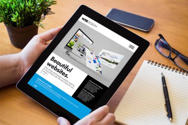 Ağaç bir çalışma masasının üzerinde web sitesi inşa cihazı tutan bir adamın elleri. Tüm ekran grafikleri uydurma.