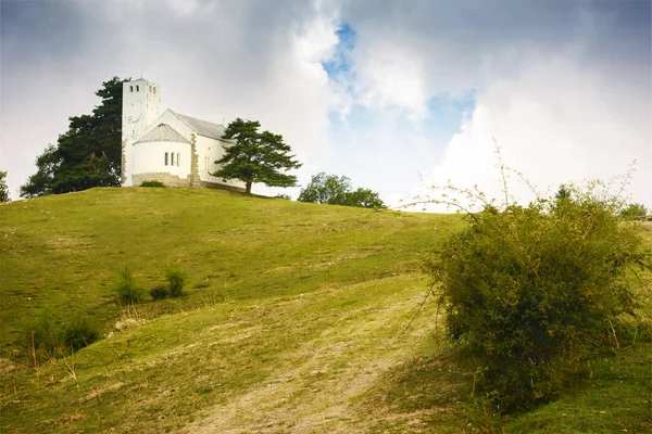 Yalnız kilise bir dağ tepe üstündeki. Yeşil çayır ağaçları ile. Italy.Cloudy gökyüzü manzara.
