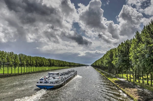 Inland barge on the long straight tree-lined Amsterdam-Rhine canal just south of Amsterdam on a day in summer with dramatic cloud formations