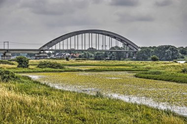 Culemborg, The Netherlands, June 30, 2017: View across the green landscape of the floodplains towards the steel arch of the railway bridge clipart