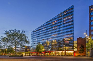 Rotterdam, Hollanda, 8 Mayıs 2019: gün batımından sonra mavi saat içinde cam zemin üzerinde restoranlar ile konut Şehir binasının görünüşte paperthin cam cephe