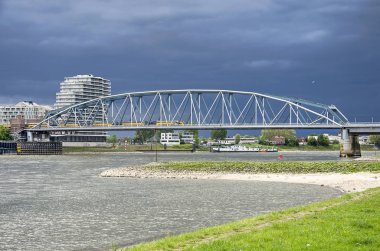 Nijmegen, Hollanda, 25 Nisan 2019: Sarı Hollanda Demiryolları treni ile demiryolu ve bisiklet köprüsüne doğru Waal nehri kumlu ve çimenli kıyısından görünümü