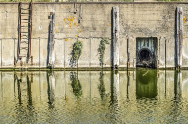 Bitişik kanala yansıyan çelik merdiven, ahşap konstrüksiyon, bitki örtüsü ve kanalizasyon çıkışı ile eski beton rıhtım duvarı