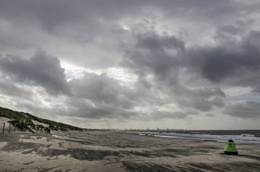 Fırtınalı bir günde Monster, Hollanda'da plaj manzarası, yeşil şamandıra şeklinde çöp kutusu, birkaç kişi, dramatik bir gökyüzü ve uzakta Rotterdam liman endüstrisi