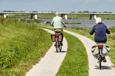 Kampen, Hollanda, 27 Temmuz 2020: Yeni Reevediep nehir kanalı boyunca beton bir yolda bisiklet süren yaşlı çift