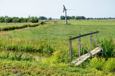 Hollanda, Schipluiden yakınlarındaki polder boyunca yürüyorum. Dar bir ahşap köprü, çelik bir yel değirmeni ve uzakta bir kilise kulesi var.