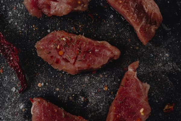 Stücke rohes Schweinemedaillons-Steak mit Gewürzen, Salz und Pfeffer auf dunklem Stabhintergrund im rustikalen Stil, Ansicht von oben — Stockfoto