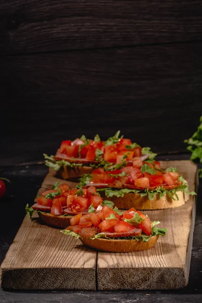 Вкусные итальянские томатные закуски, или брускетта, на ломтиках поджаренного багета, украшенного листьями салата, ветчиной, нарезанными помидорами и соусом, закрытыми на деревянной доске — стоковое фото
