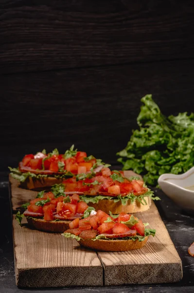 Saboroso saboroso tomate aperitivos italianos, ou bruschetta, em fatias de baguete torrado decorado com folhas de salada, presunto, tomate picado e molho, close-up em uma tábua de madeira — Fotografia de Stock