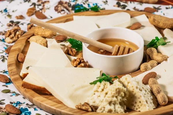 Assorted cheeses on round wooden board plate served with white wine Guda cheese, cheese grated bark of oak, hard cheese slices, walnuts, grapes, crackers, honey, sulguni, light wood background — Stock Photo, Image