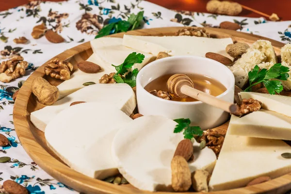 Assorted cheeses on round wooden board plate served with white wine Guda cheese, cheese grated bark of oak, hard cheese slices, walnuts, grapes, crackers, honey, sulguni, light wood background — Stock Photo, Image