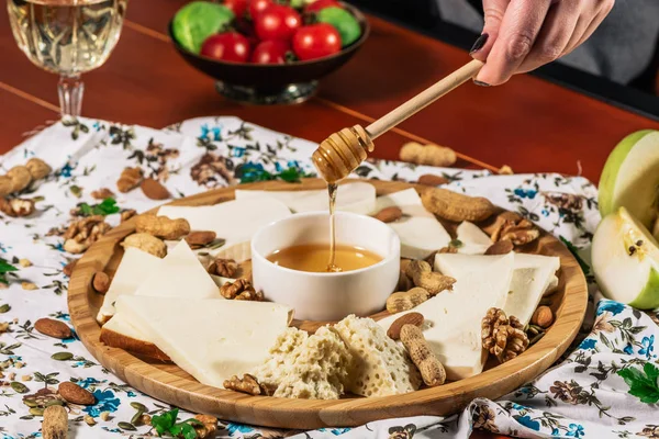 Cheese plate and chef hand wich pours honey: Sulguni, Guda, cheddar, gouda, camembert, and other served with white wine, honey and nuts on wooden board on light background. Tasty appetizers. — Stock Photo, Image