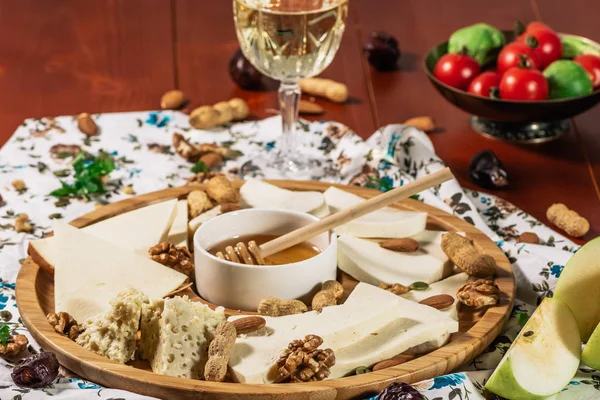 Assorted cheeses on round wooden board plate served with white wine Guda cheese, cheese grated bark of oak, hard cheese slices, walnuts, grapes, crackers, honey, sulguni, light wood background — Stock Photo, Image