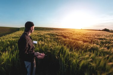 Şapkalı genç tarımcı gün batımında yeşil bir tarla arazisinde not alıyor.