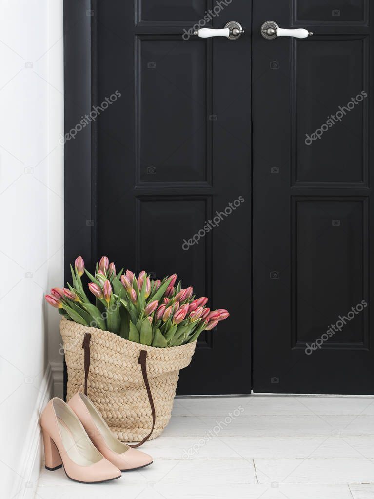 Nude shoes and straw bag with bunch of fresh pink tulips on floor near black door