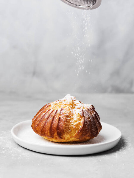 Французская профитроль с заварным кремом, посыпанным и порошковым сахаром на белой тарелке на сером фоне
