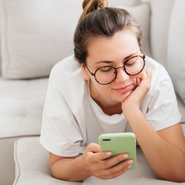 Gülümseyen genç kadın gri kanepede smartphone kullanarak görünümünü kapat