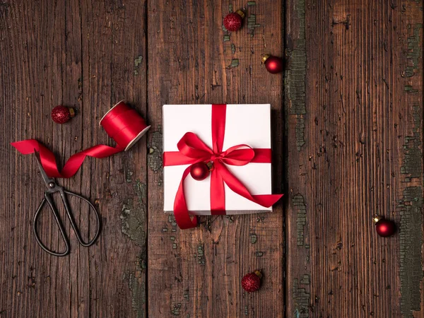 white box decorated red ribbon and red christmas balls with scissors and wrapping tape on vintage wooden background