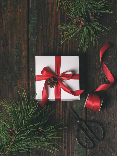 белый ящик украшен красной лентой с сосновыми ветвями и шишками с ножницами и оберточной лентой на винтажном деревянном фоне, вид сверху
 