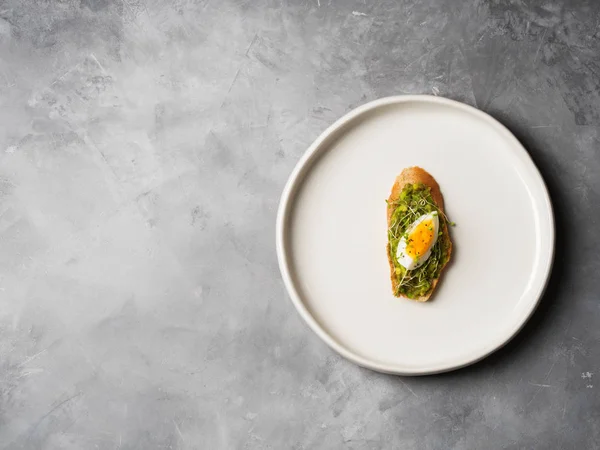 Whole-grain toast plated avocado with eggs and fresh mini herbs with sunflower seeds and sprinkled black salt served on ceramic white plate over grey concrete background, top view, Diet food concept, gluten-free