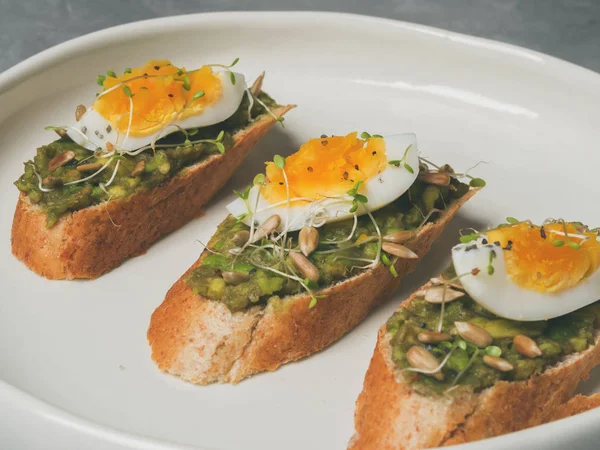 Whole-grain toasts plated avocado with eggs and fresh mini herbs with sunflower seeds and sprinkled black salt served on ceramic white plate over grey concrete background, top view, Diet food concept, gluten-free